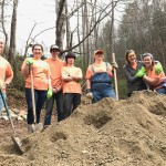 Group of people in orange shirts standing with shovels next to a pile of dirt