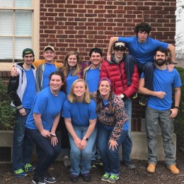 Group of students in blue shirts posing in front of a brick building
