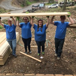 Four people in blue shirts holding a wooden board on a construction site
