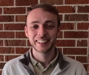 A young man with short light brown hair and a trimmed beard smiles at the camera. He is wearing a light gray jacket over a beige collared shirt and is standing in front of a red brick wall